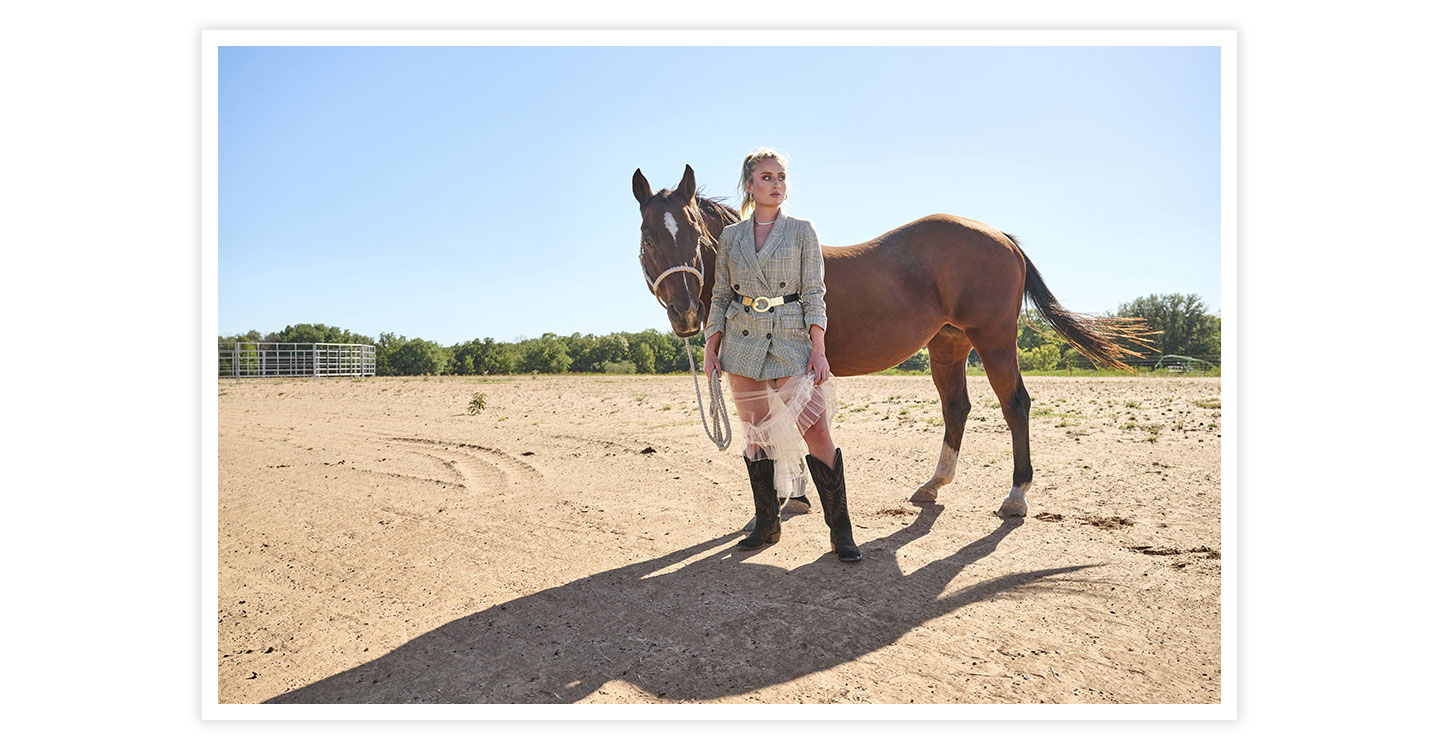 A woman wearing Clara 15” Western Boot in black holding a horse lead rope with a horse, in a dirt field.